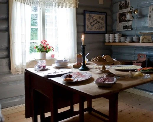 Tradisjonelt kjøkken med trebenk, blomsterpotte på vinduskarmen, borddekkning med stearinlys, bøker og kjøkkenutstyr.