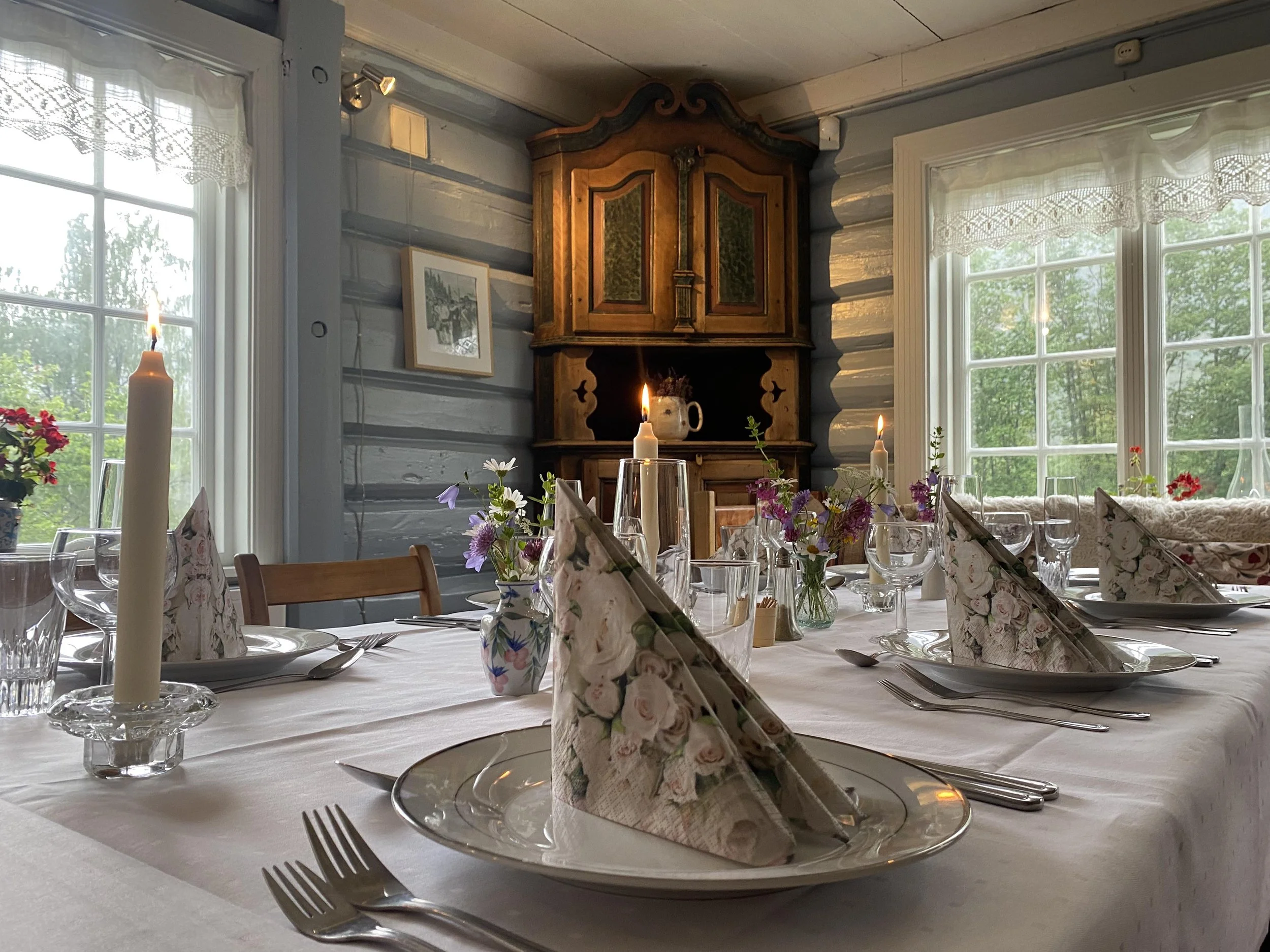Et bord dekket til middag med blomsteroppsats, stearinlys, glass, tallerkener, bestikk og pyntede servietter i en rustikk stue med store vinduer og gamle bilderammer.