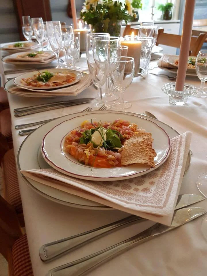Festbord dekket med hvit duk, flere tallerkener med forrett, vin- og vannglass, lysestaker og telys, blomster midt på bordet, samt bestikk.