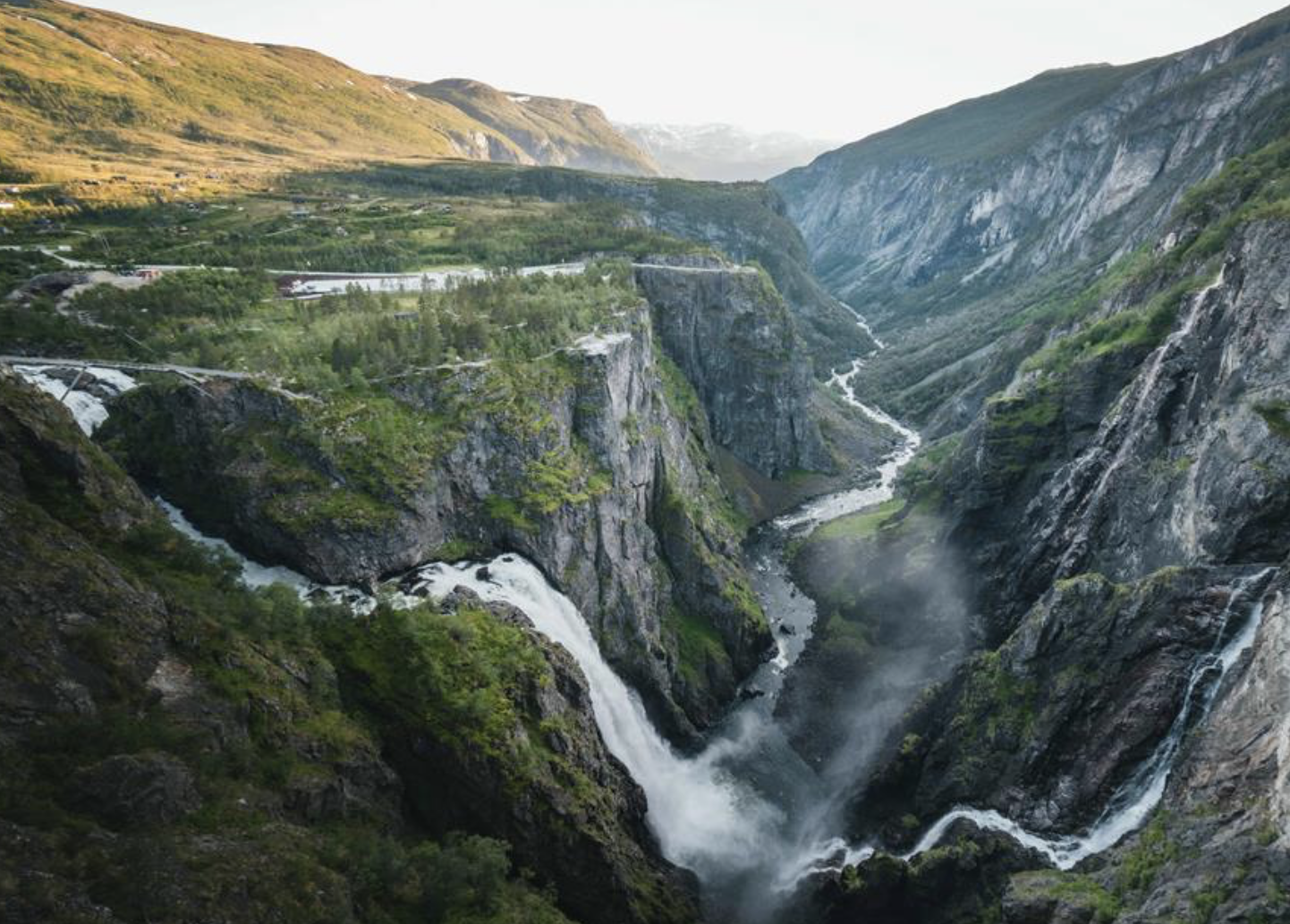 En dyp og bratt kløft med fossefall som fosser ned, omgitt av grønne fjellsider og spredte bygninger i bakgrunnen.