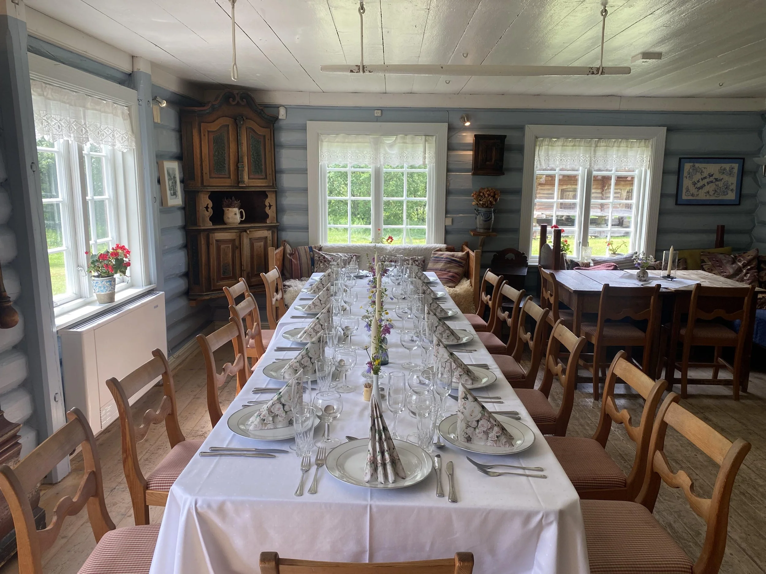 Innendørs bilder av et langbord dekket til fest med hvit duk, blomster og stearinlys, omgitt av stoler og benker, i et rom med store vinduer og tradisjonell dekorasjon.