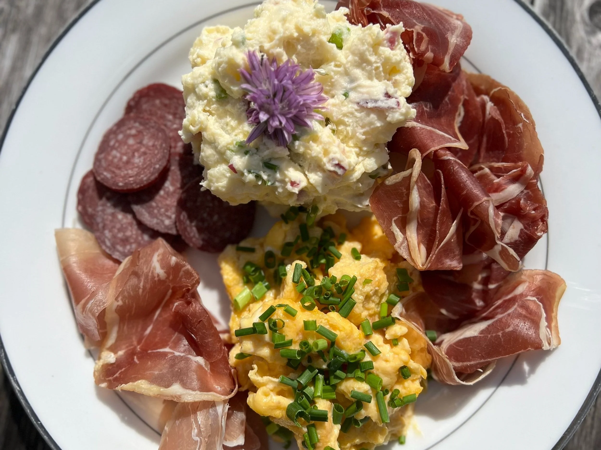 Spekemattallerken med spekeskinke, fenalår, sognemorr, hjemmelaget potetsalat og salat fra hagen. Midt på tallerkenen er det en klatt med eggerøre pyntet med en lilla blomst og hakket grønn løk.