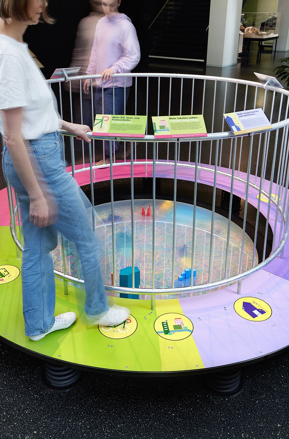 Two people stand on the outer raised ring of the circular exhibit. Holding onto the handrail and operating the foot pedals, they watch as the CO₂ fog dissipates, gradually revealing the city at the center of the exhibit.