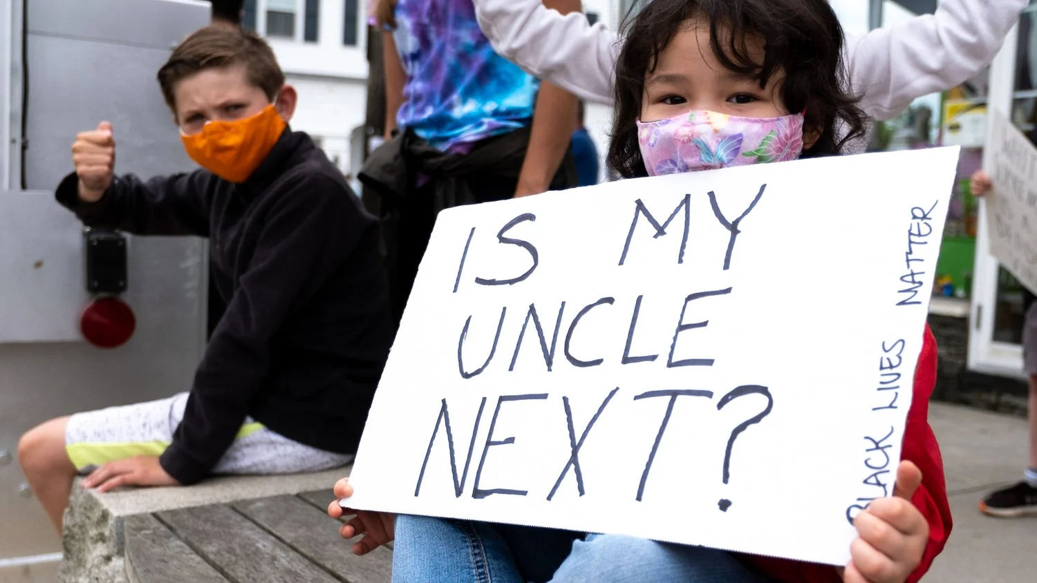 Masked girl with an “Is my uncle next?” sign beside a masked boy
