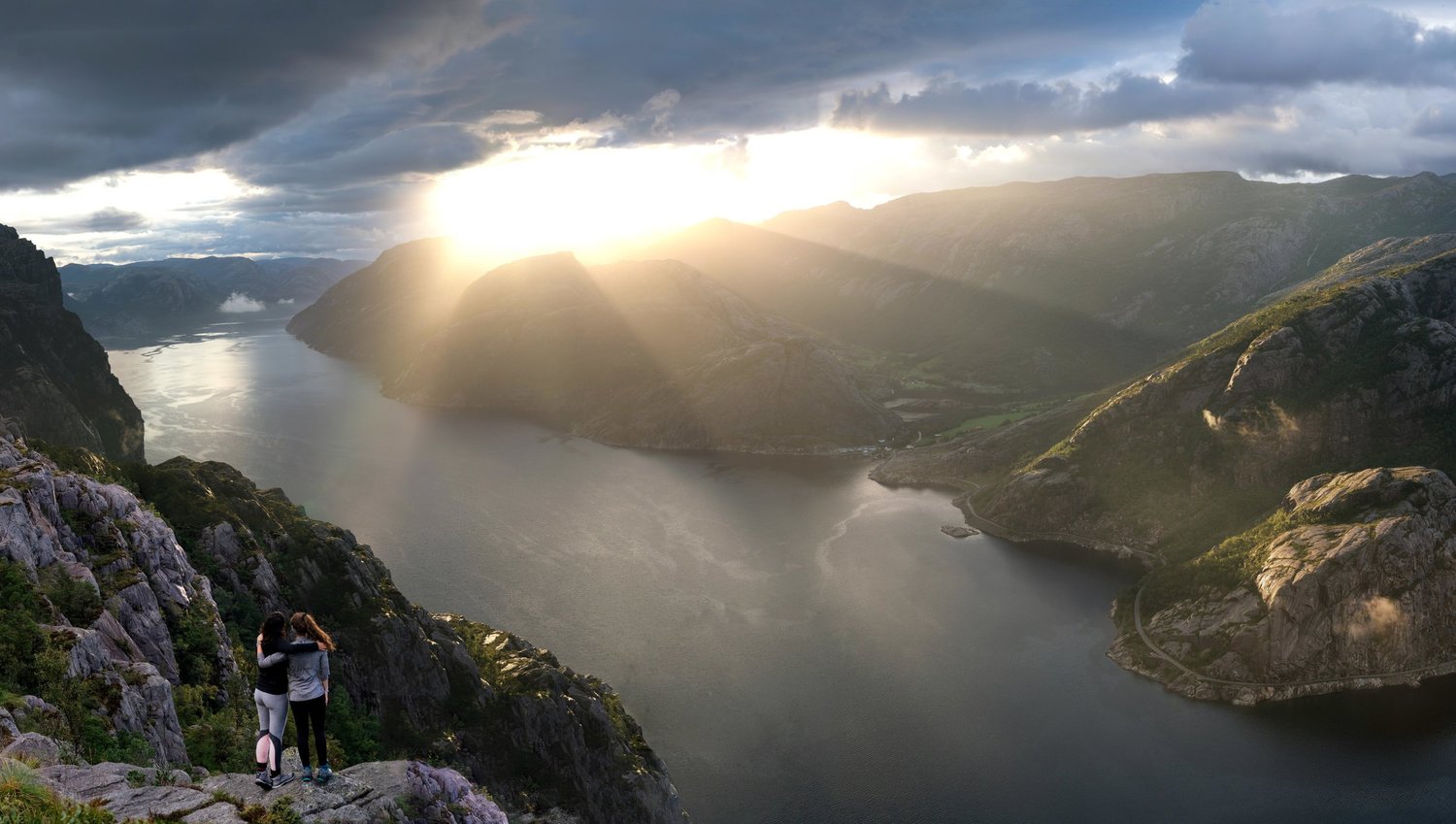 Sunbeams breaking over a Norwegian fjord, hikers on the cliff edge