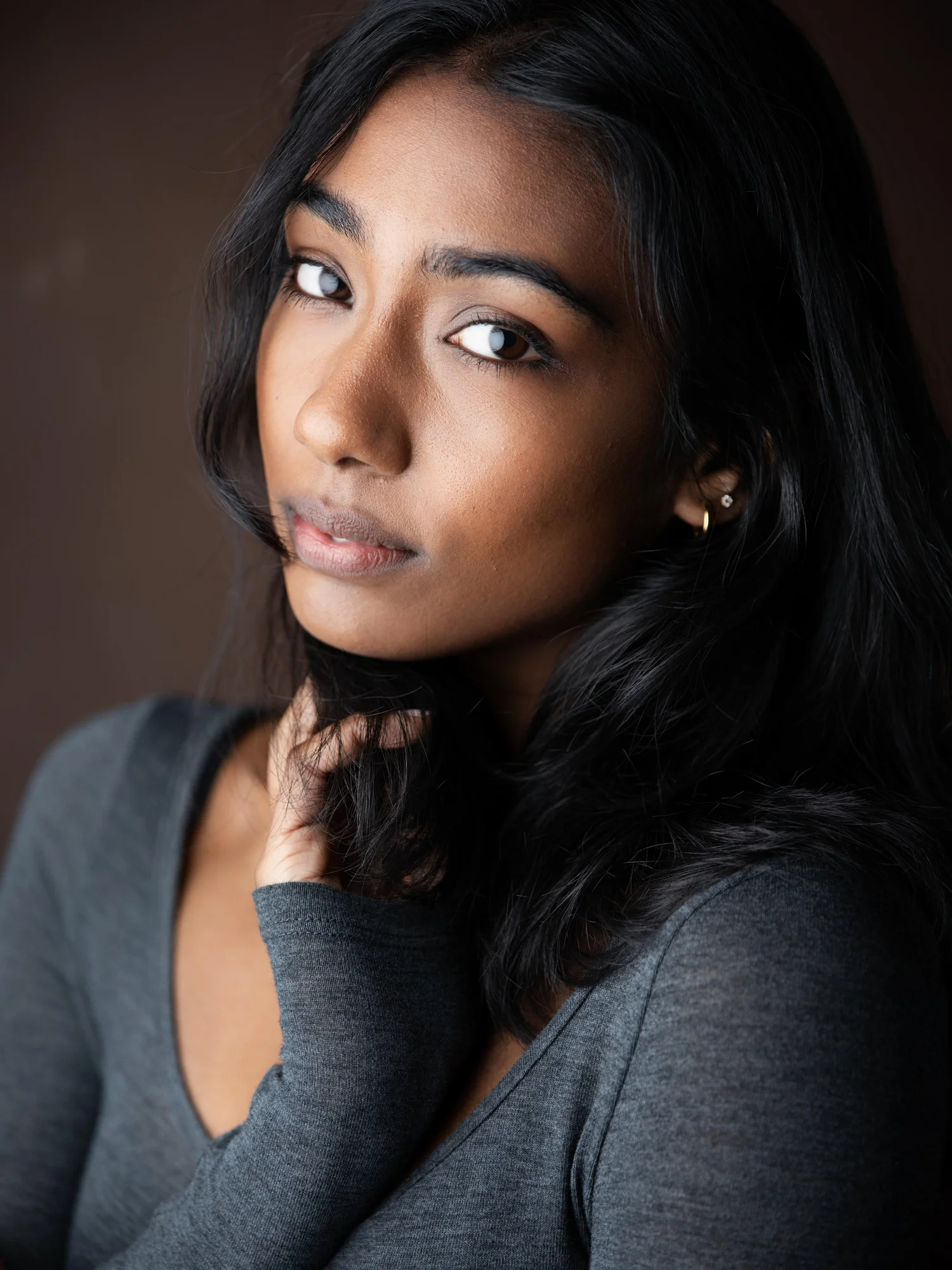 Woman with long dark hair resting her fingertips on her cheek against a dark backdrop