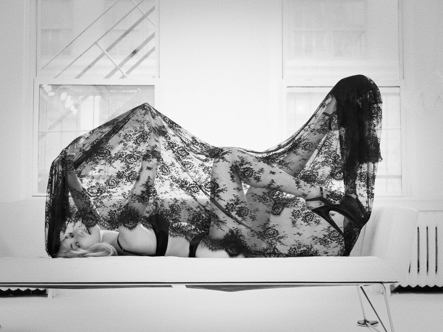 Model reclining on a white sofa beneath a sheet of black lace, heels raised