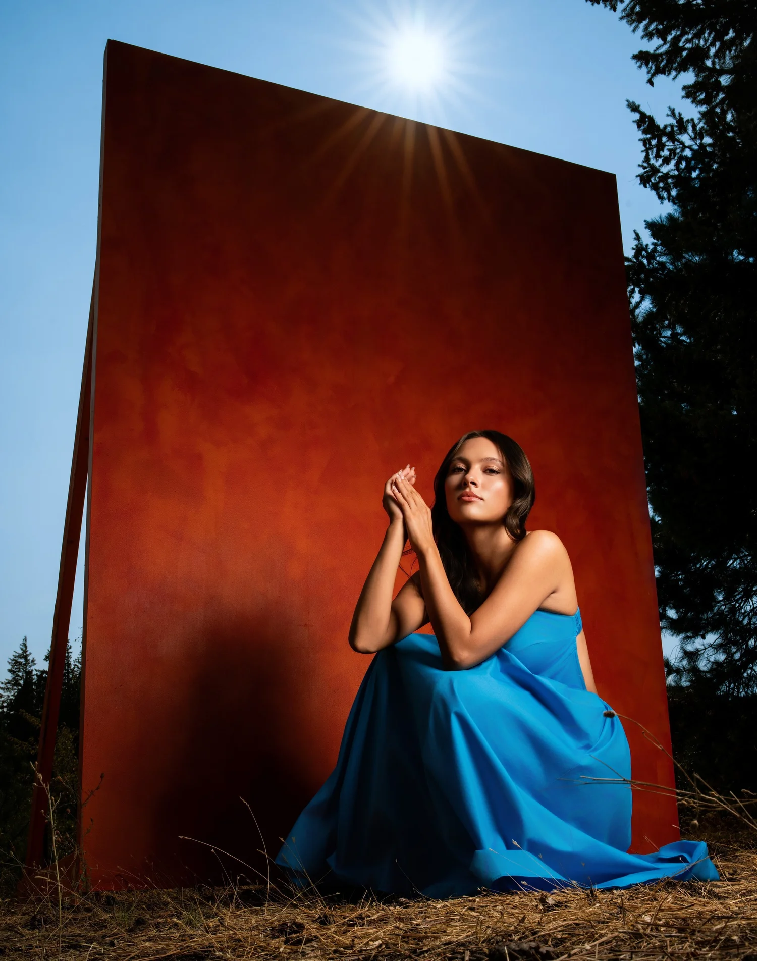 Woman in a blue dress seated before a towering rust-red steel wall, the sun flaring over its edge