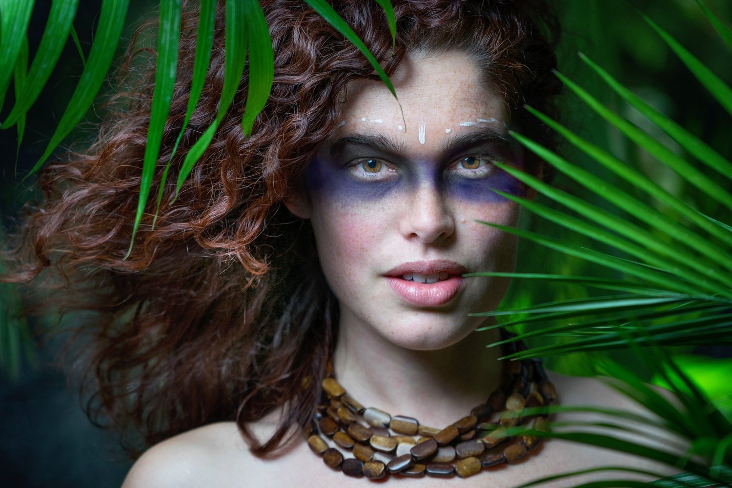 Woman with curly hair and blue eye makeup framed by palm fronds, wearing a wooden bead necklace