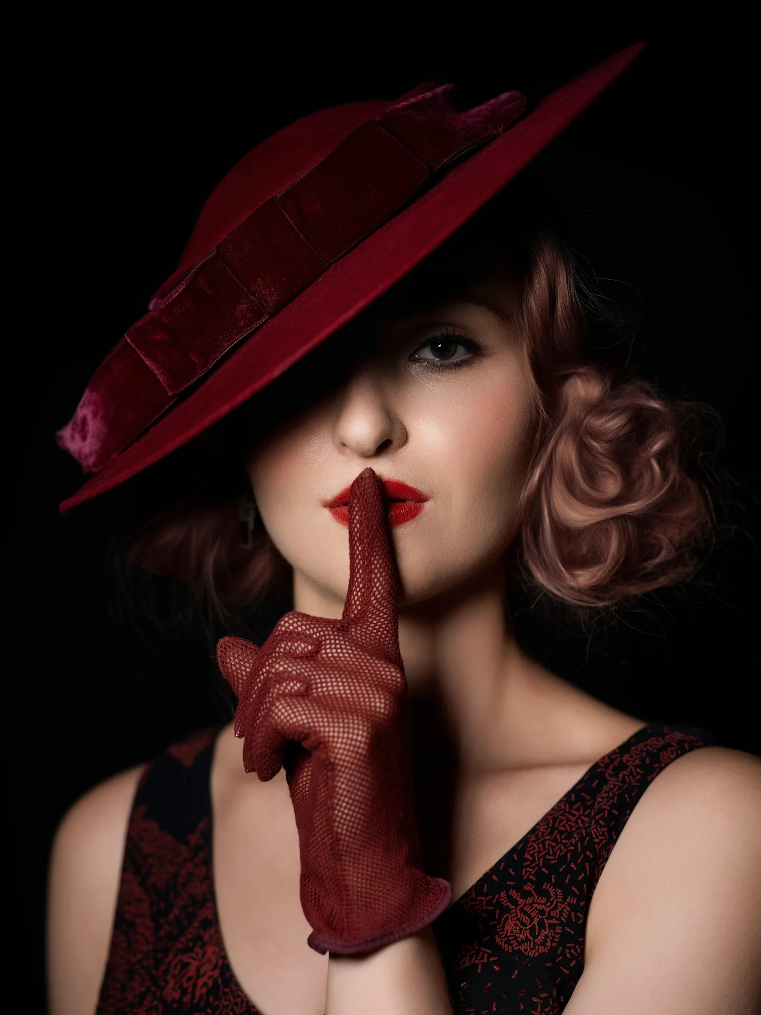 Woman in a red hat and red gloves holding a finger to her lips against a dark background
