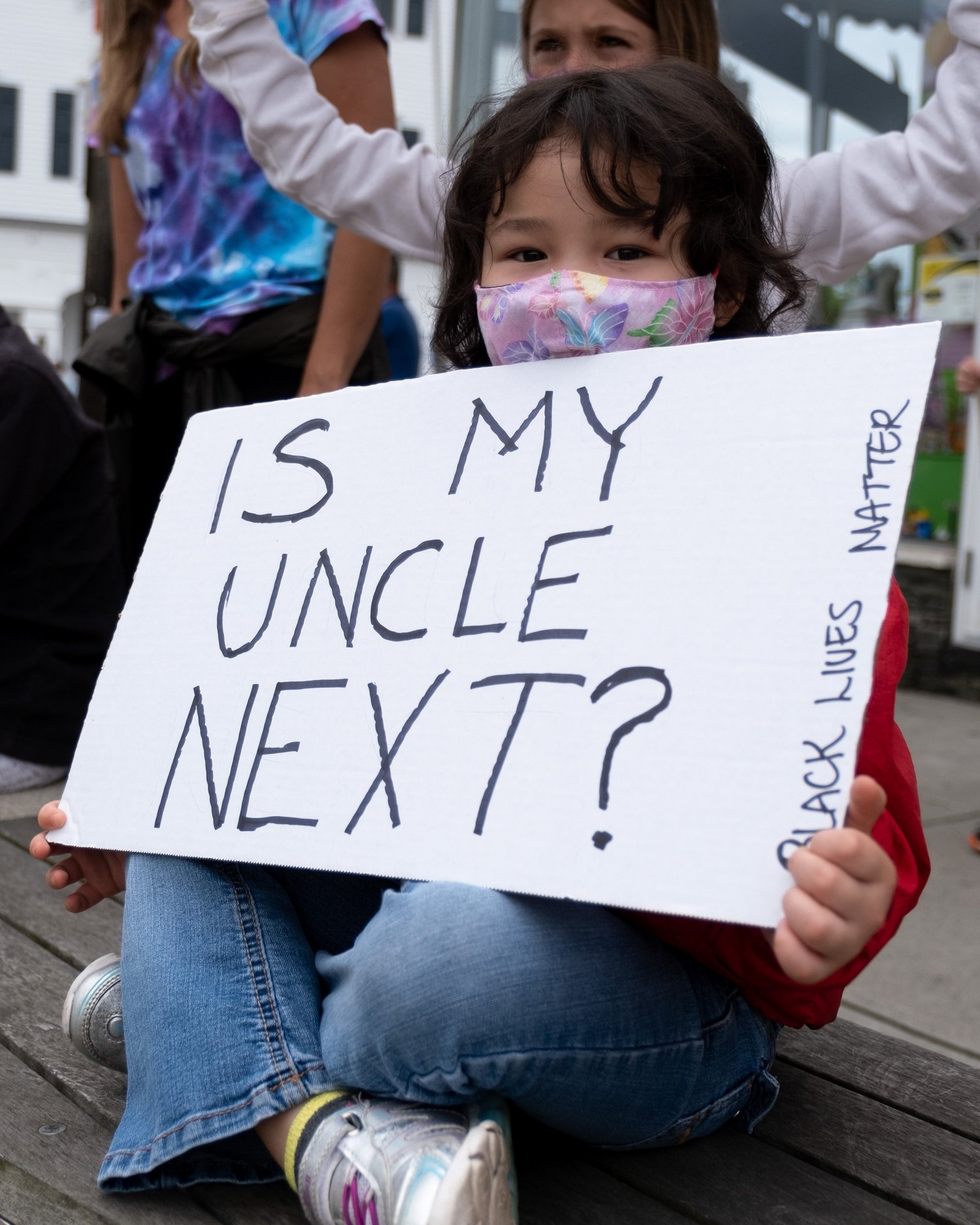 Masked girl holding a sign that asks “Is my uncle next?”
