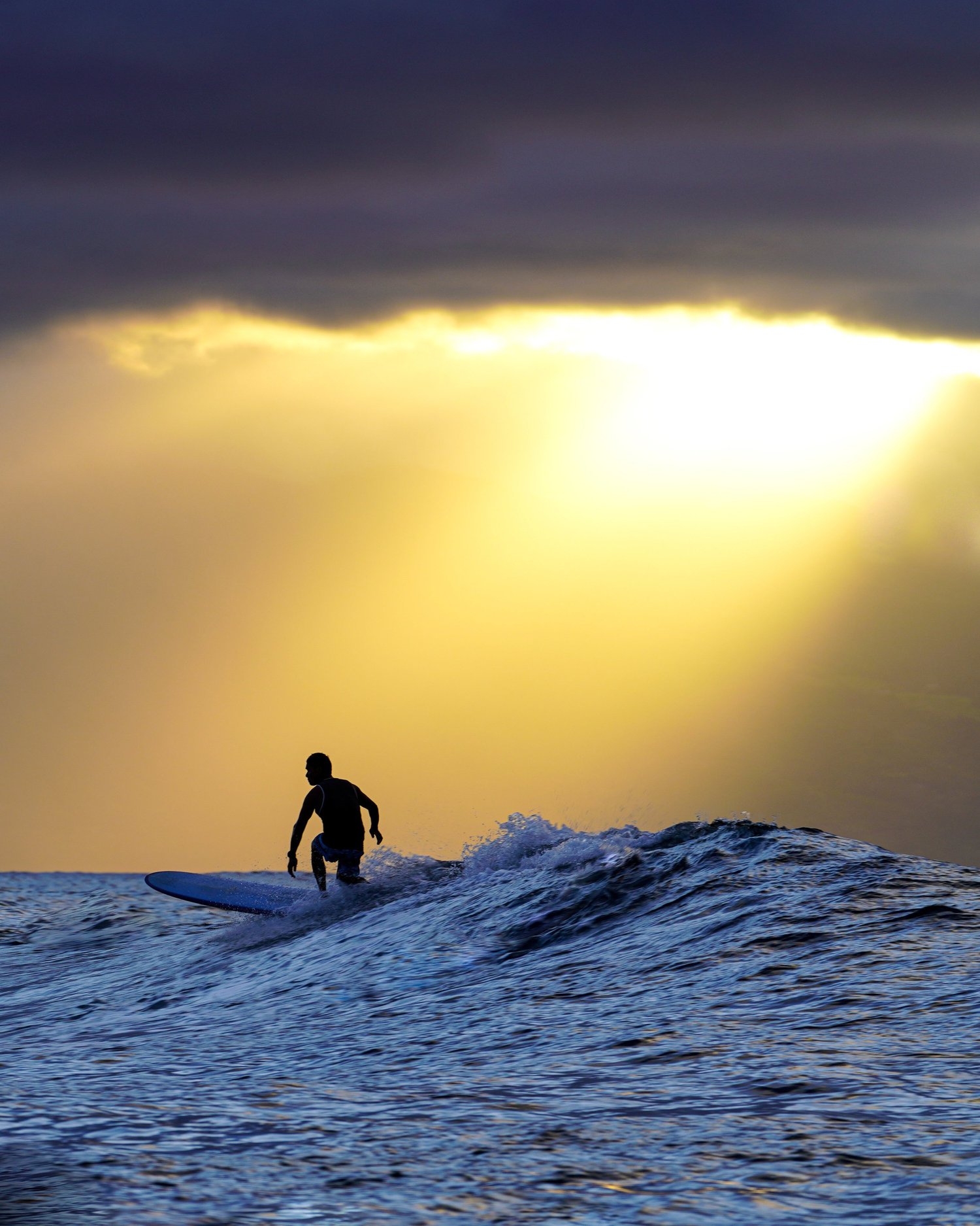 A silhouetted surfer on a wave beneath light breaking through clouds
