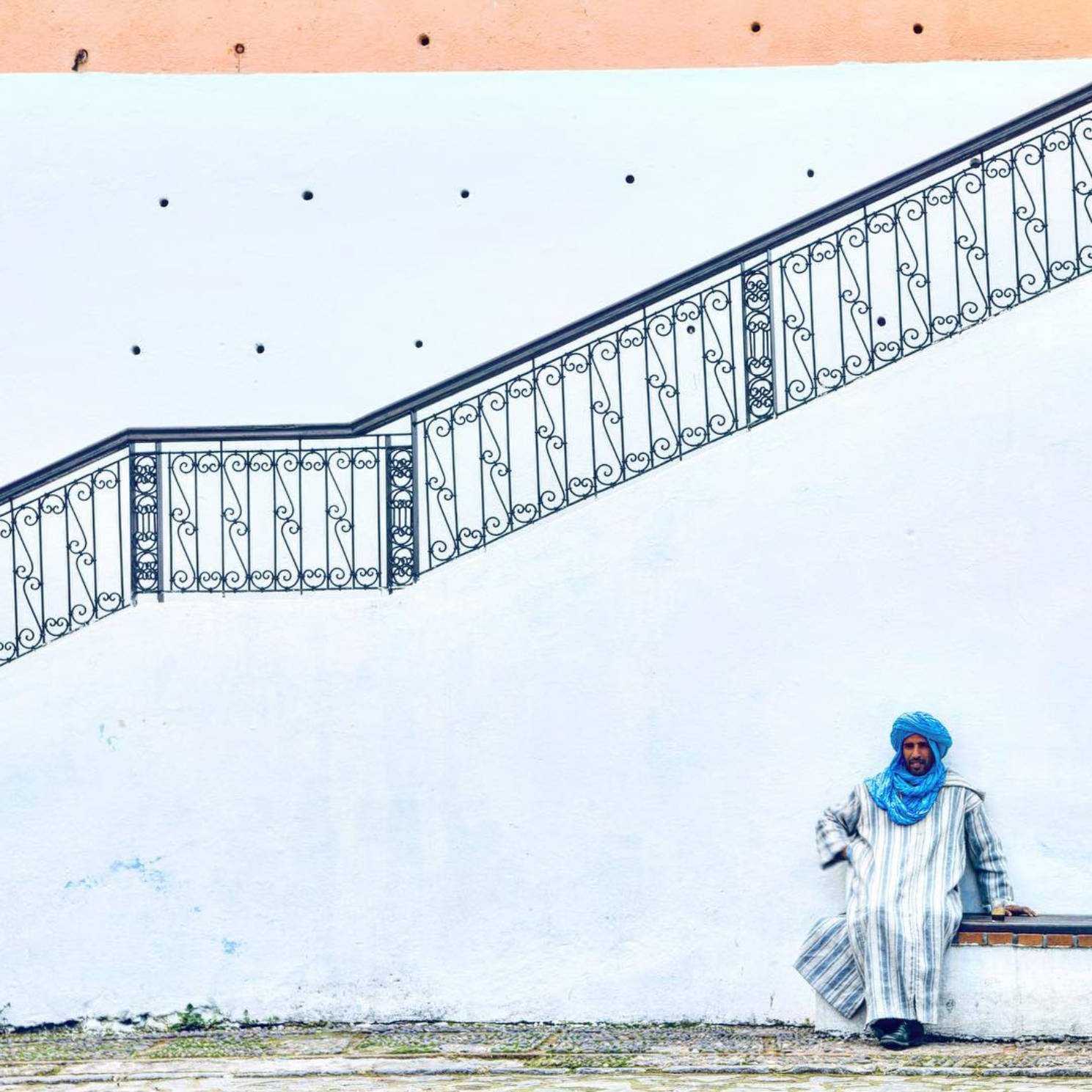 Man in a blue robe seated on white stairs beneath an iron railing