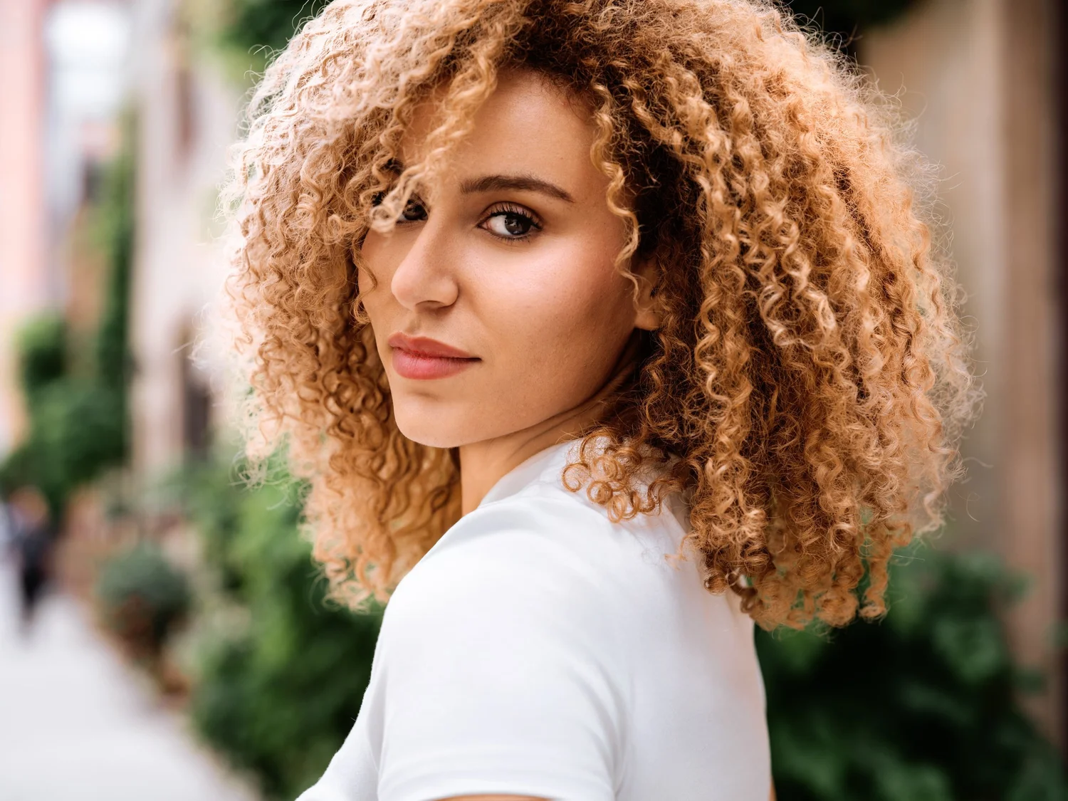 Woman with voluminous curly hair in a white T-shirt on a leafy street
