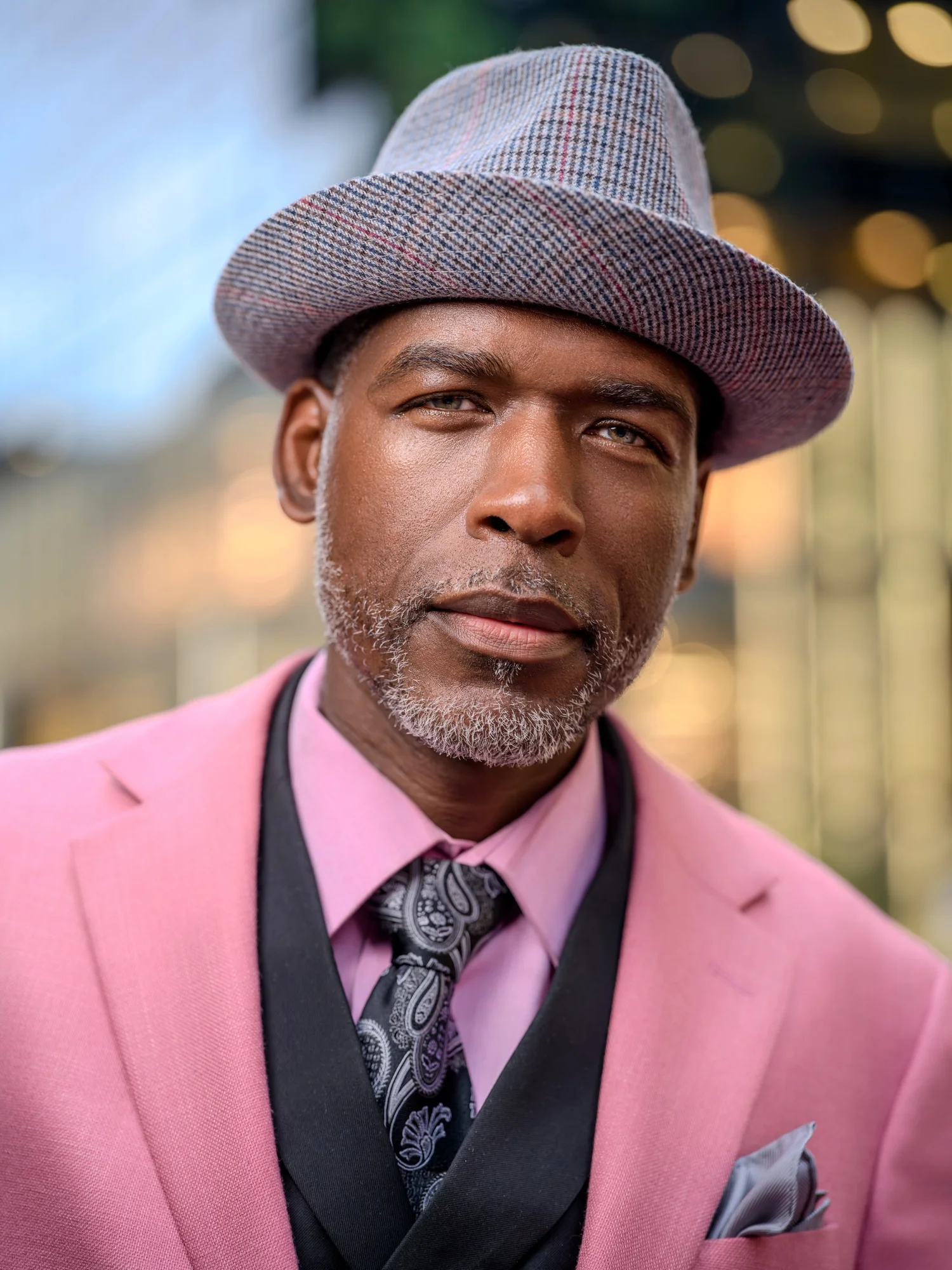 Jason Jamal in a pink suit, patterned tie and gray fedora, city lights behind him