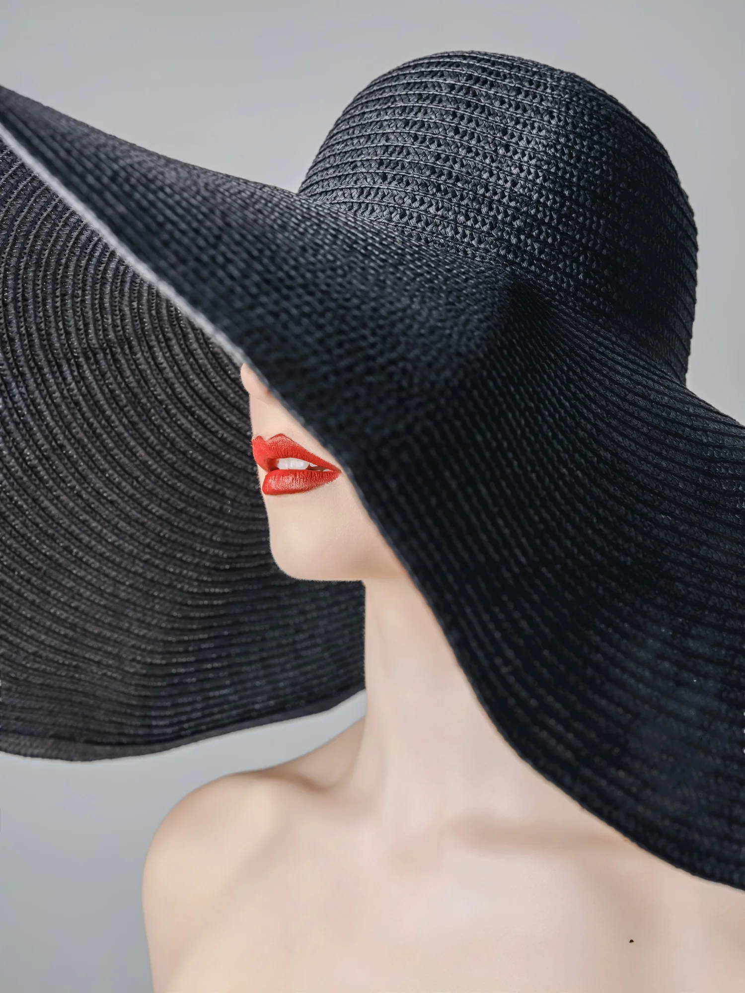 Woman in a wide black sun hat that hides her eyes, red lips, pale gray backdrop