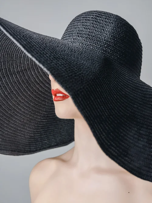 Woman in a wide black sun hat that hides her eyes, red lips, pale gray backdrop