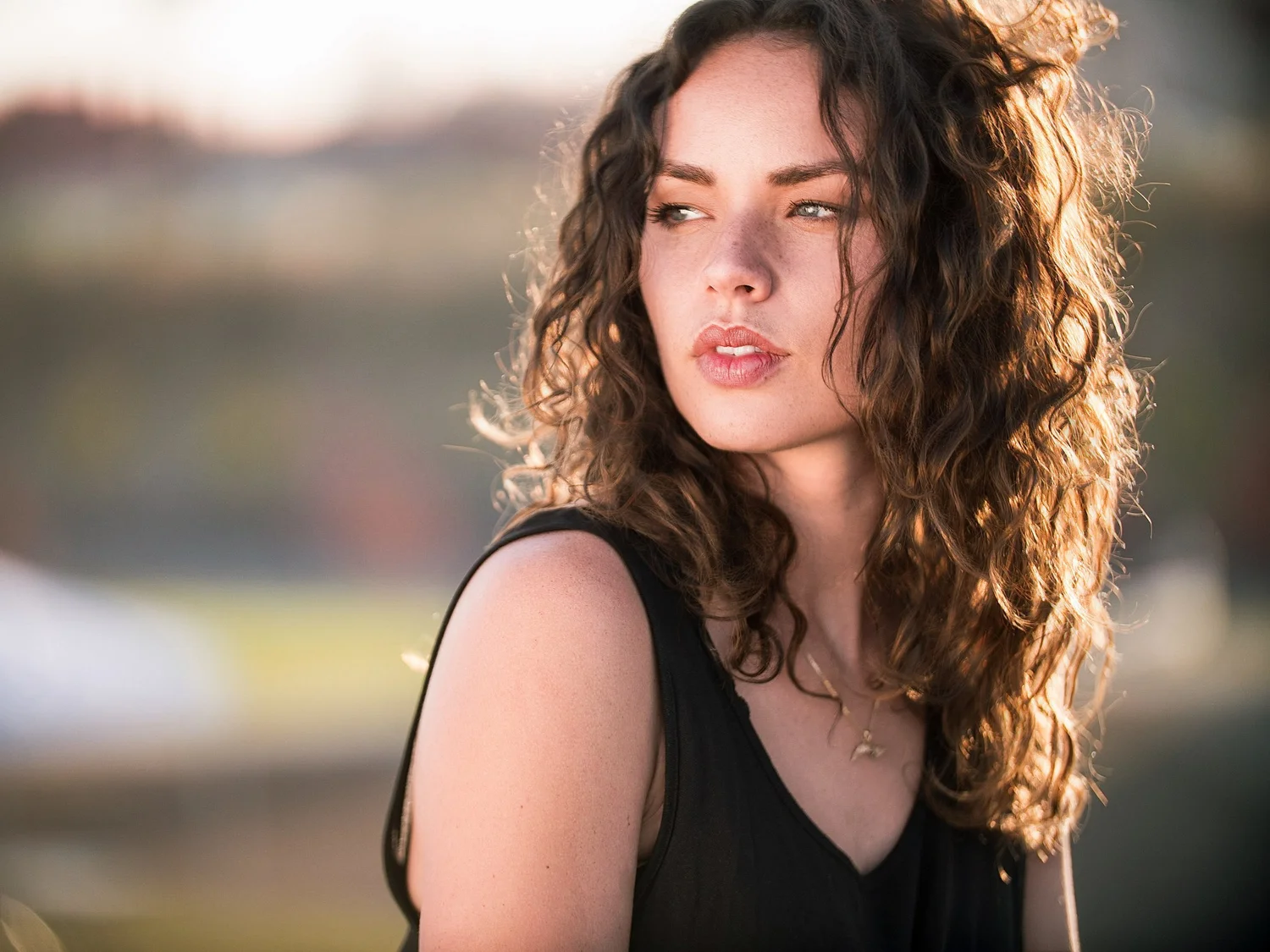 Woman with dark curly hair in a black top looking off to the side, warm light behind her