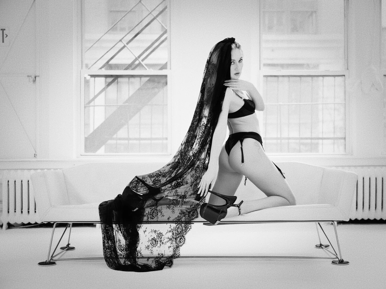 Model in black lingerie kneeling on a white sofa in window light, a long black lace veil trailing to the floor