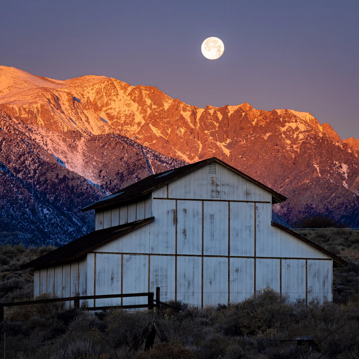 White barn beneath snowy mountains lit by sunset, the full moon above
