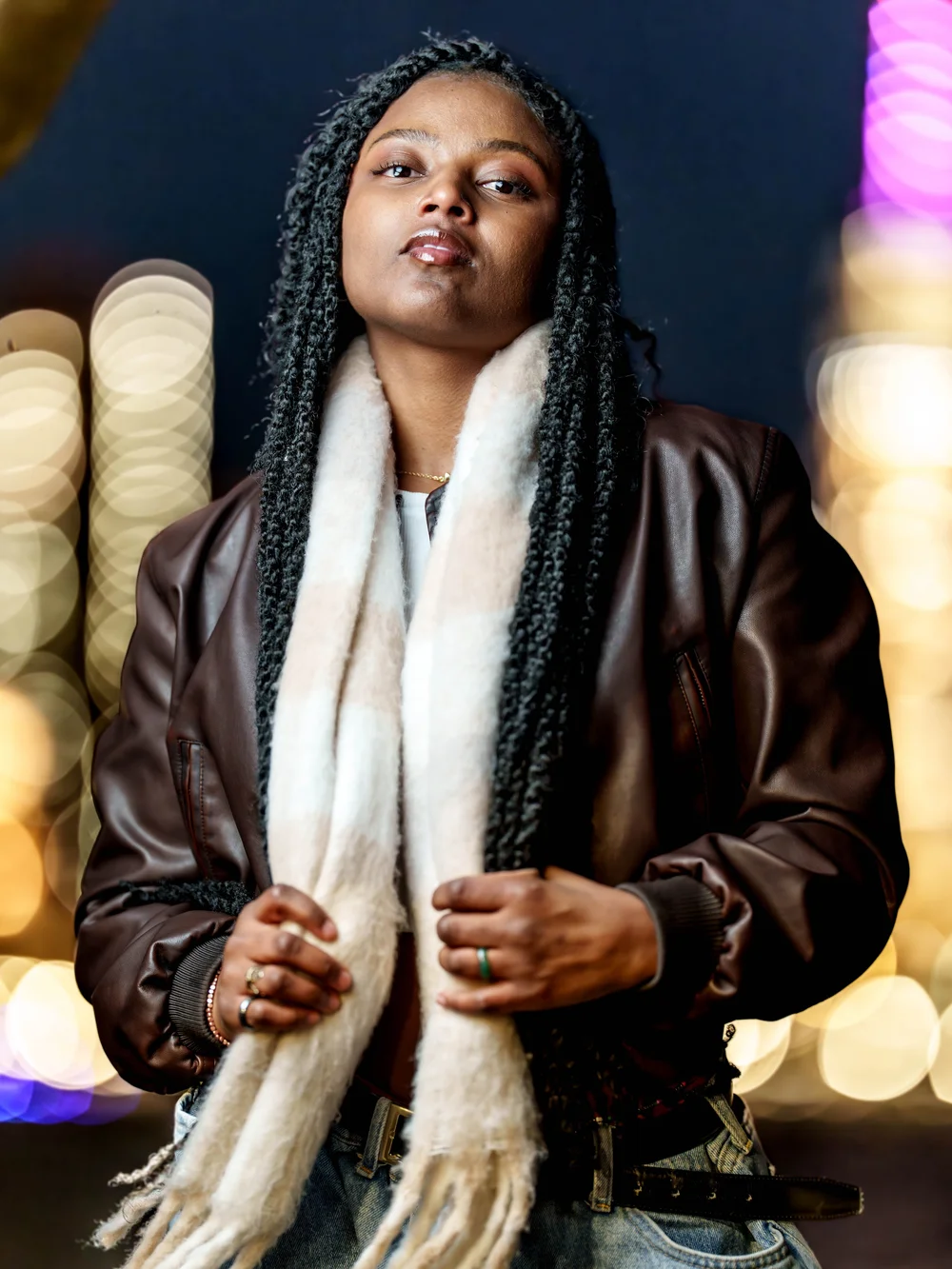 Woman with long braids in a leather jacket and white scarf, city lights behind her