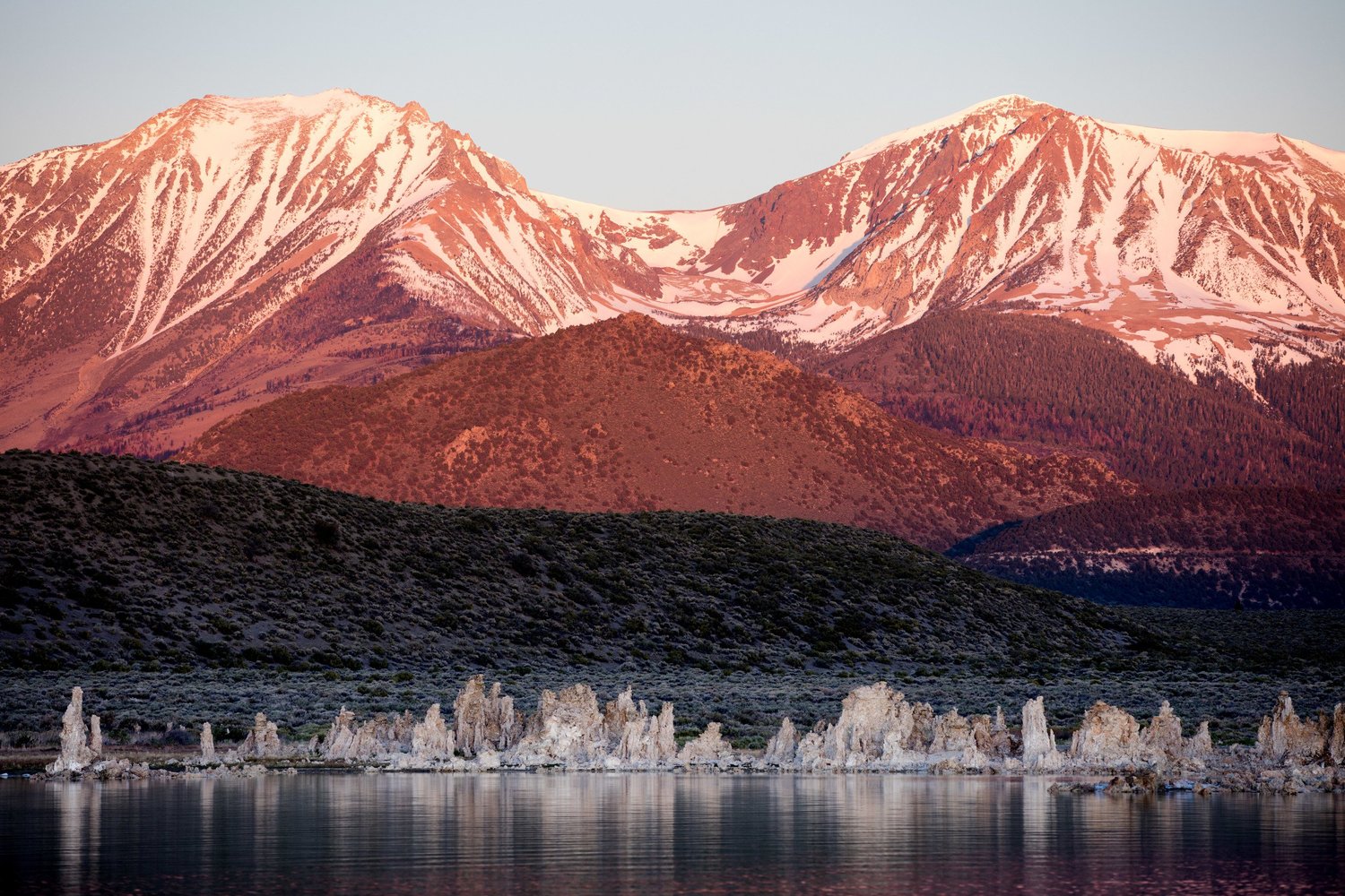 Snowy peaks glowing pink at dawn above a still lake lined with tufa towers
