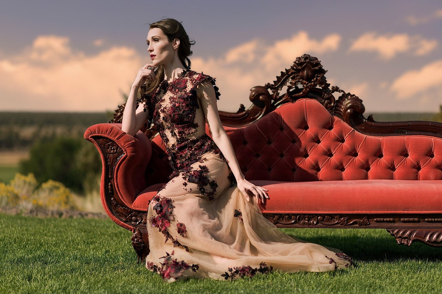 Woman in a floral gown seated on a red Victorian sofa on a lawn at sunset