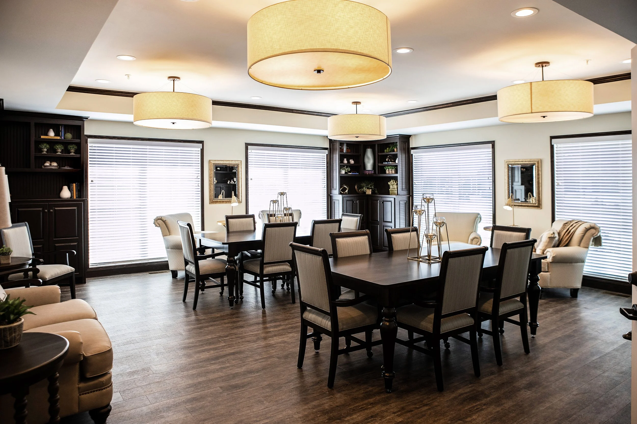 Elegant dining room with large wooden tables, upholstered chairs, and overhead lighting. The room has wood flooring, bookshelves, and large windows with blinds.