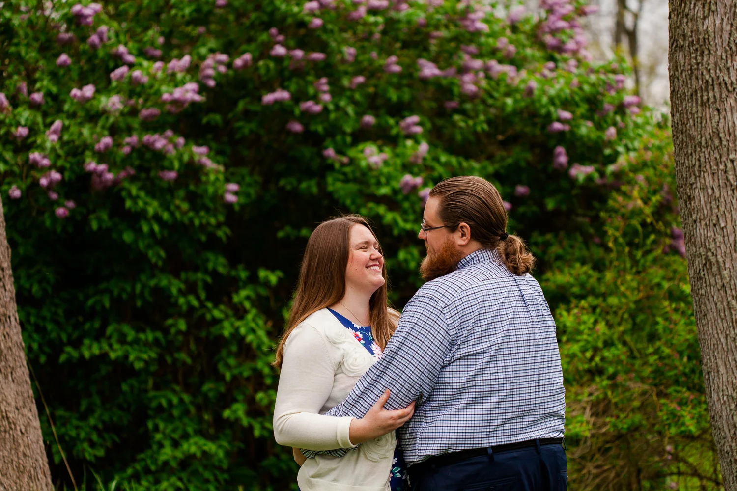 Woman smiles at man in front of lilac bush during engagement shoot.