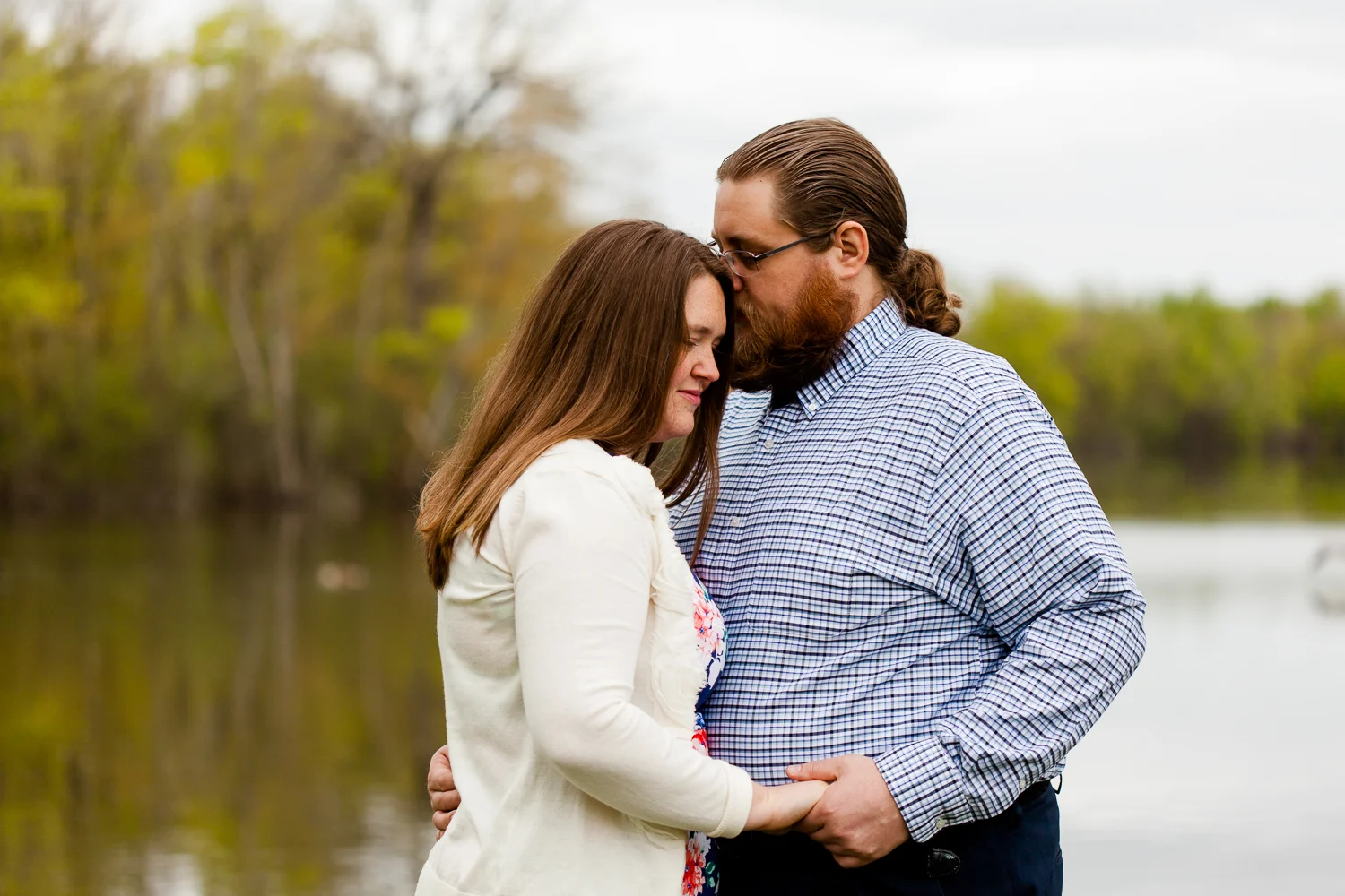 A man kisses the forhead of a woman in a white sweater during their spring engagement session. They are standing in front of a lake.