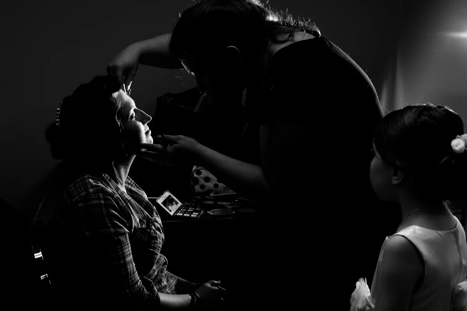 Flower girl watches bride as she gets her make up done. A black and white image with dramatic lighting.