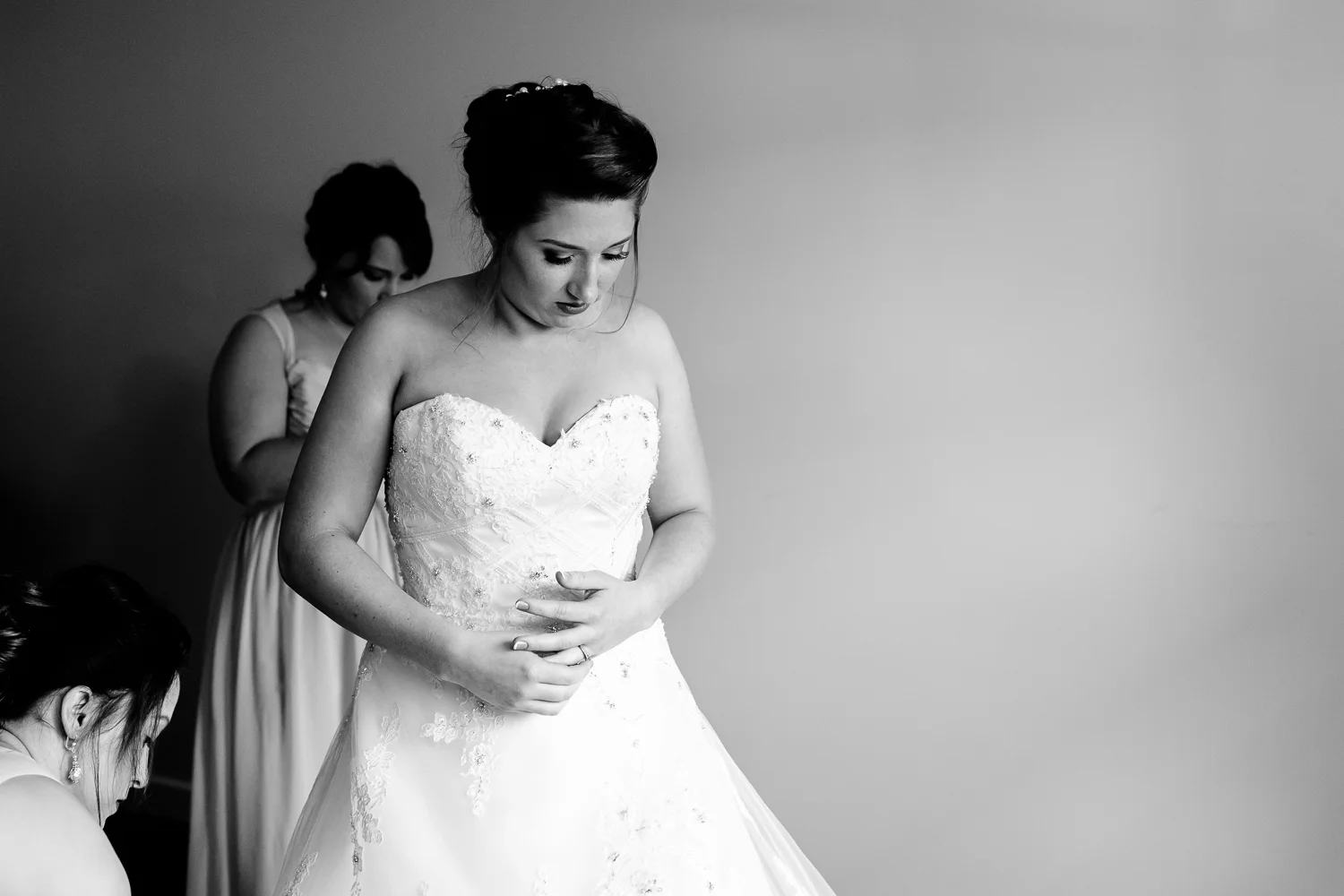Black and white image of a bride putting on her wedding gown. Her bridesmaids help her.