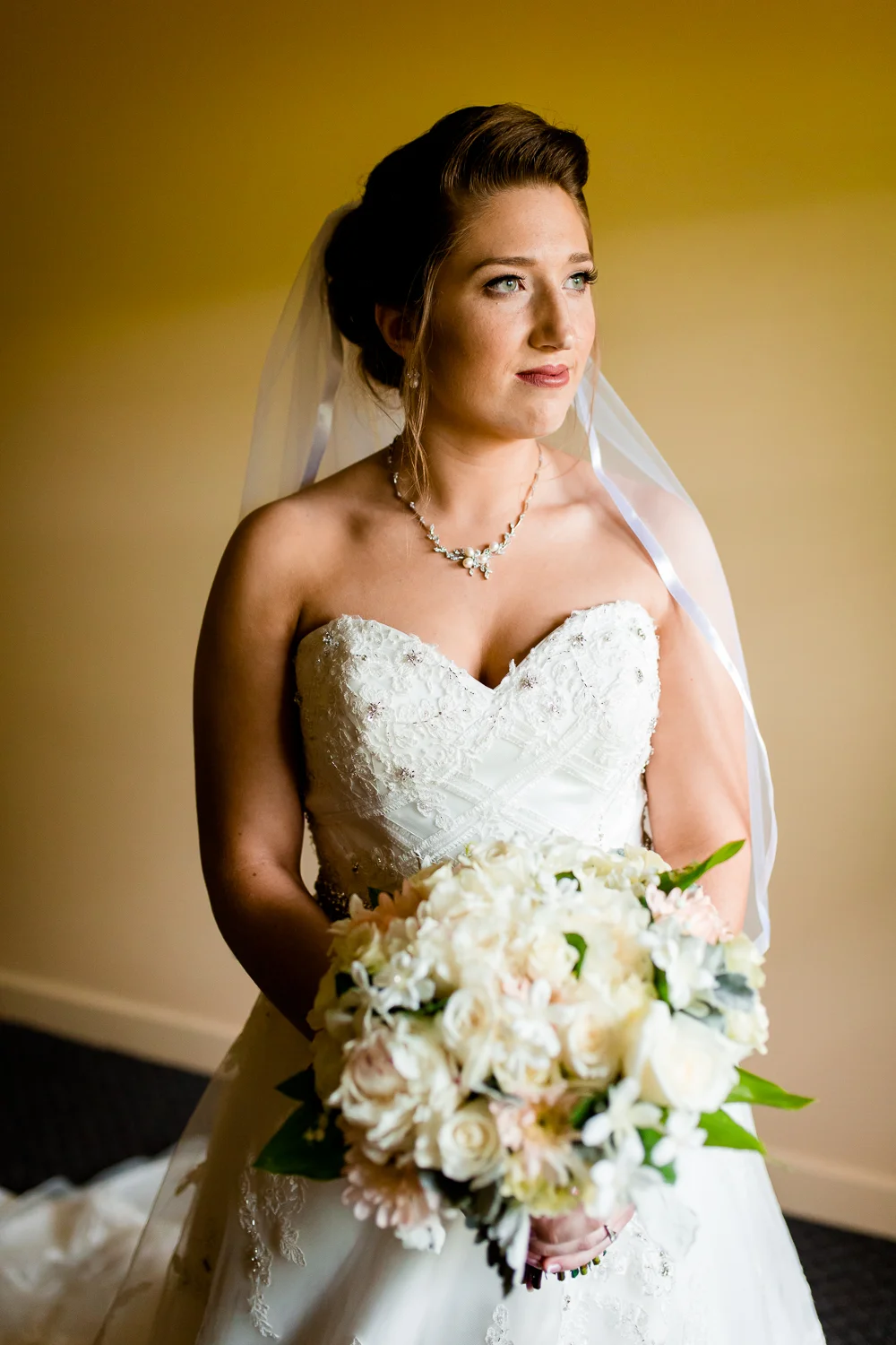 Portrait of bride holding her bouquet of flowers. She is wearing a veil and gazing out of the window. 