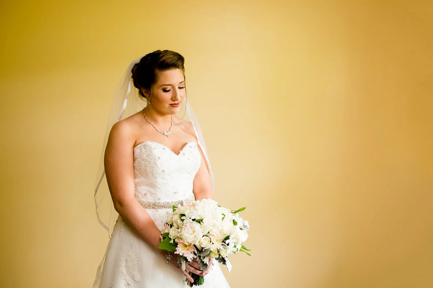 Bride looks down at her bouquet of white flowers. She is in a white wedding gown with a veil and she is standing against a stark yellow wall.
