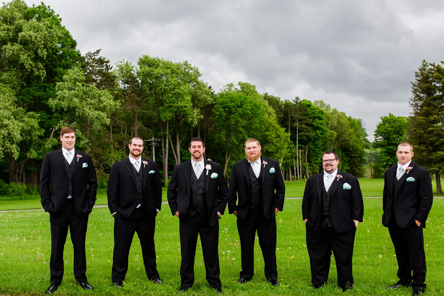 The groom and groomsmen stand in a line against a back drop of green trees. The sky is dark with clouds.