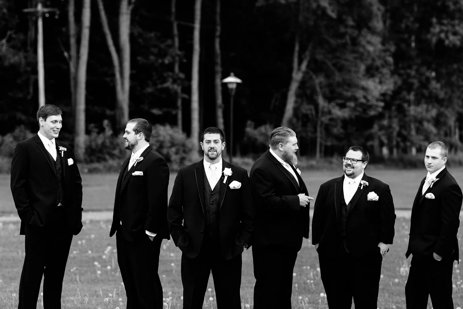 A black and white image of the groom and groomsmen. The groom looks at the camera while the groomsmens talk behind him.
