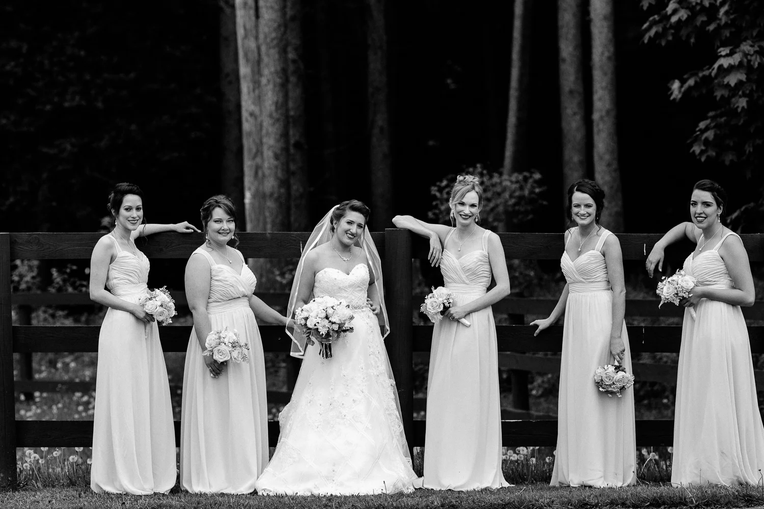Black and white image of bridesmaids and bride leaning against a fence.