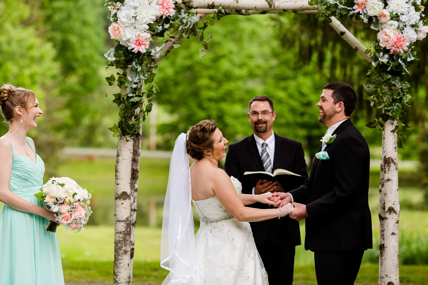 Bride and groom laugh during their wedding ceremony. They are under a birch tree arch. The maid of honor stands next to them laughing.