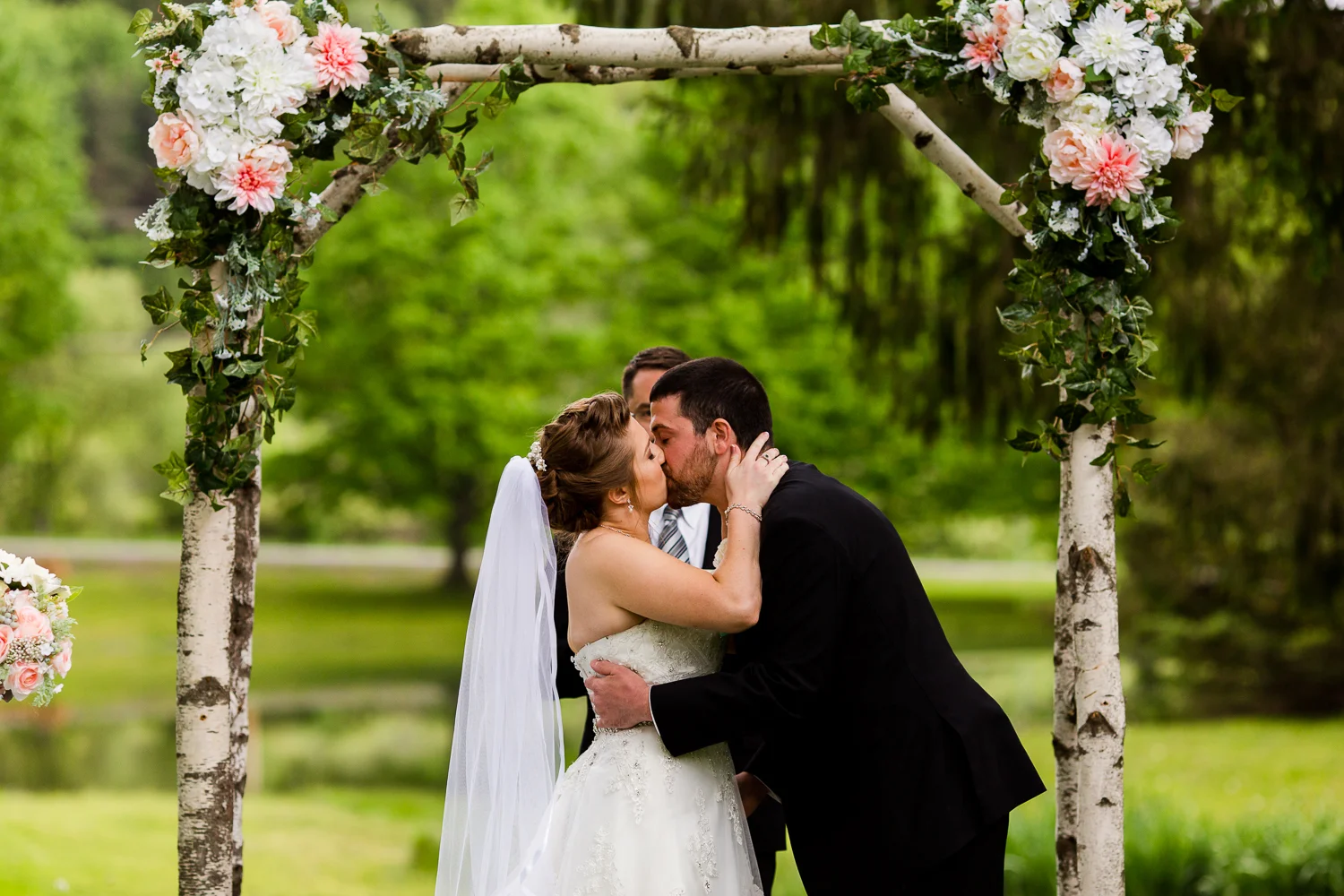 Bride and groom share their first kiss during their outdoor wedding ceremony. They are standing under a birch tree arch adorned with flowers. There is a pond in the background.