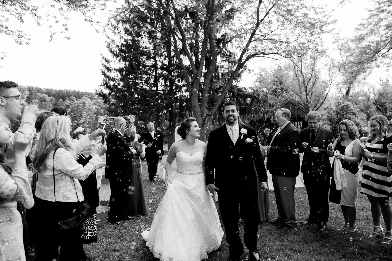 Bride and groom walk down the aisle post wedding ceremony as guests blow bubbles around them. The image is in black and white. The bride is smiling up at the groom.