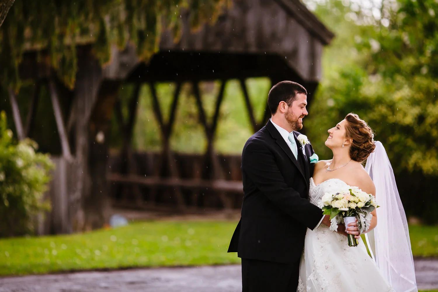 Bride and groom look at each other. They are standing in front of a covered wooden bridge.