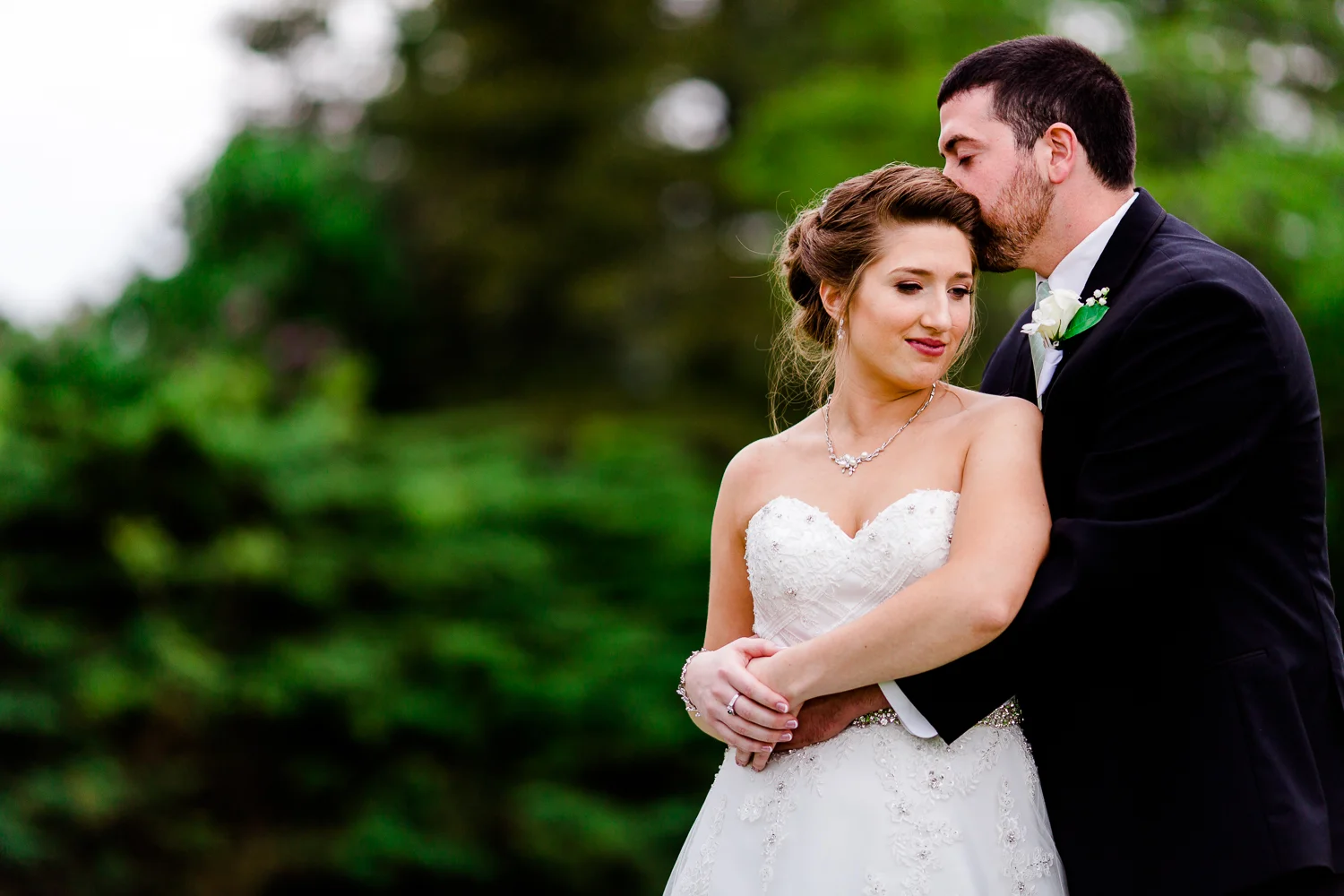Groom kisses bride on the side of the head while bride gazes off over her shoulder. There are green trees behind them.