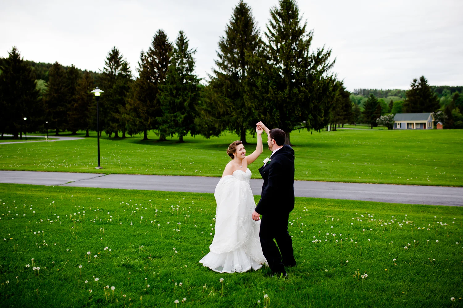 Bride and groom twirl on green grass. There are white flowers around them. Pine trees are in the distance. The bride is holding her white lace wedding gown in one hand.