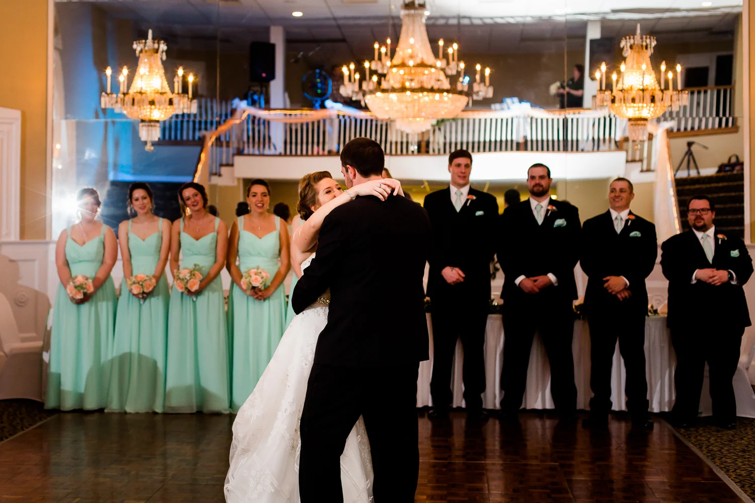 First dance of bride and groom. The wedding party looks on. There are chandeliers hanging above them.