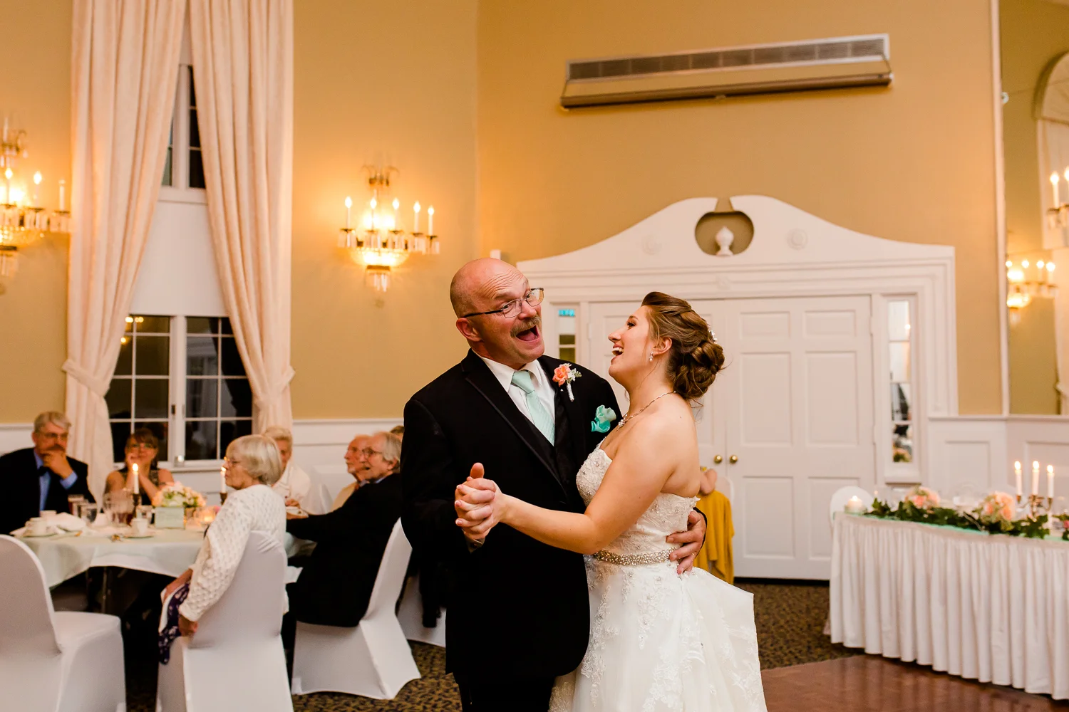 The bride and her father laugh during the father daughter dance at her wedding. They are in a large ball room.