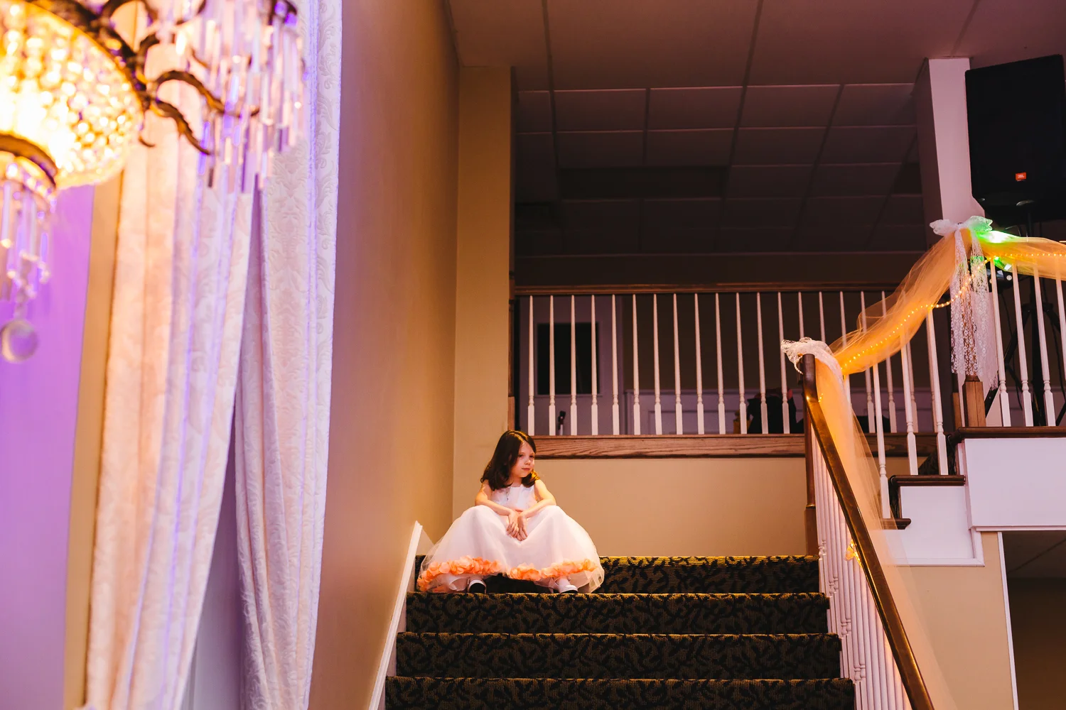 Flower girl sits on the stairs at the wedding reception. She is looking down on the dance floor.
