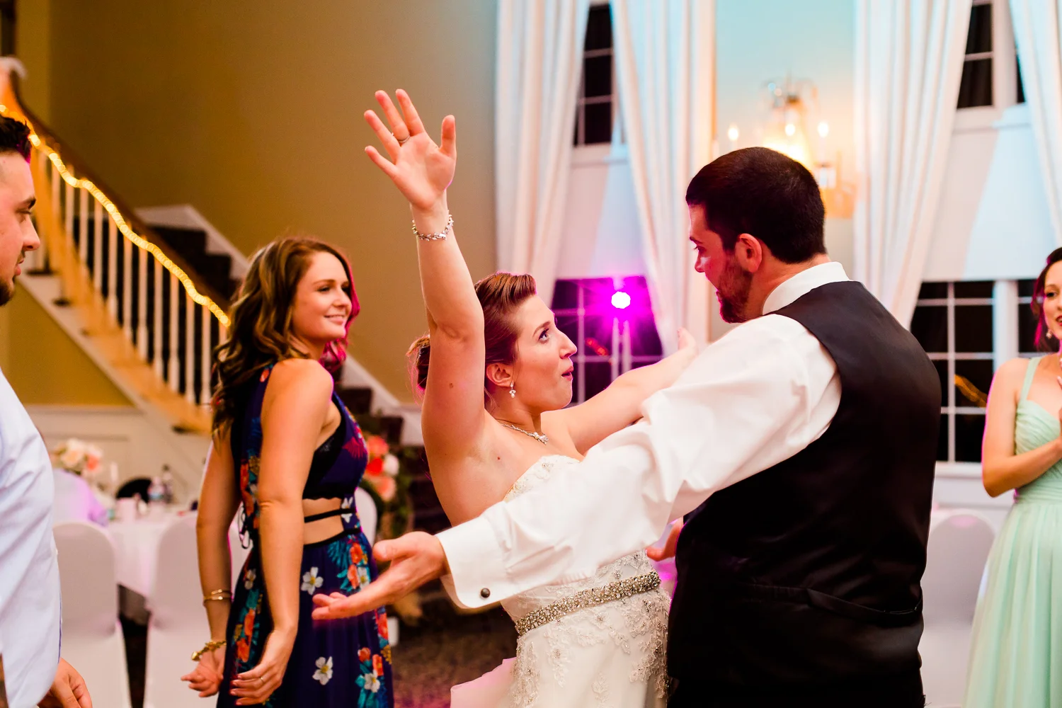 Bride and groom share a silly dance moment on at their wedding reception. The bride has her arms up in the air. Friends dance in the background.