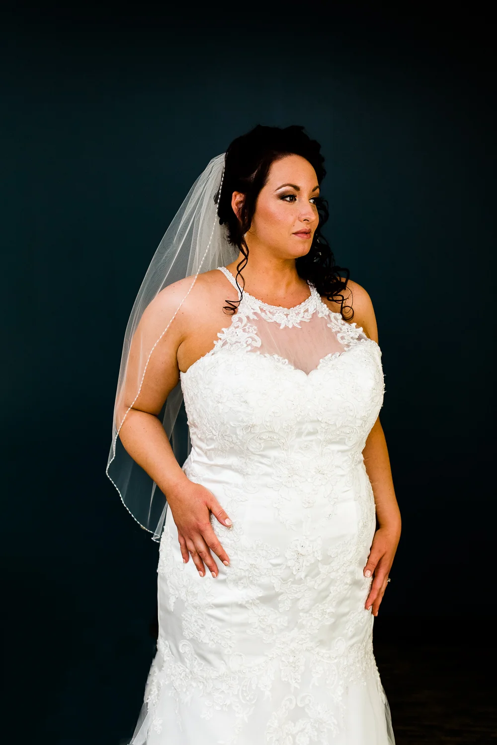 Bride in white halter gown with lacework. She is against a blue background looking off into the distance.