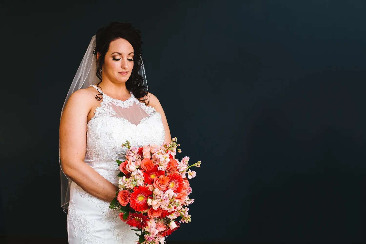 Bride looks down at giant bouquet of coral flowers. She is in a white gown. Her hair has curls. She has a veil on.