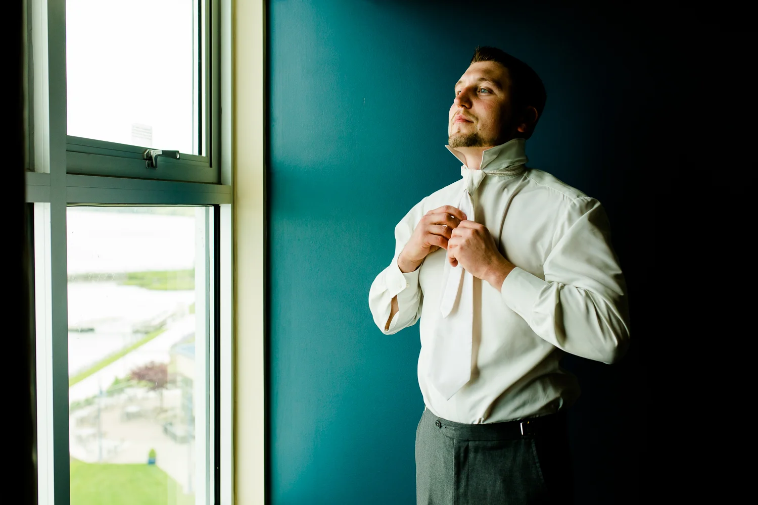 Groom adjusts tie against blue background. He is looking out of a window.
